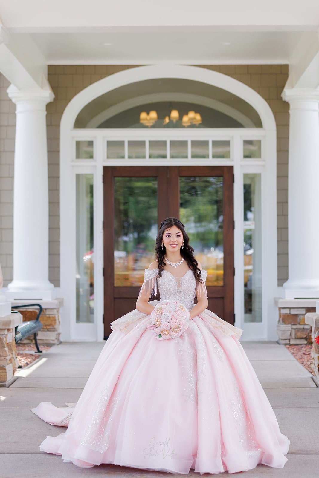 Quinceañera in a blush pink ball gown holding a rose bouquet in front of the columned entrance at Glen Oaks Country Club in West Des Moines, Iowa